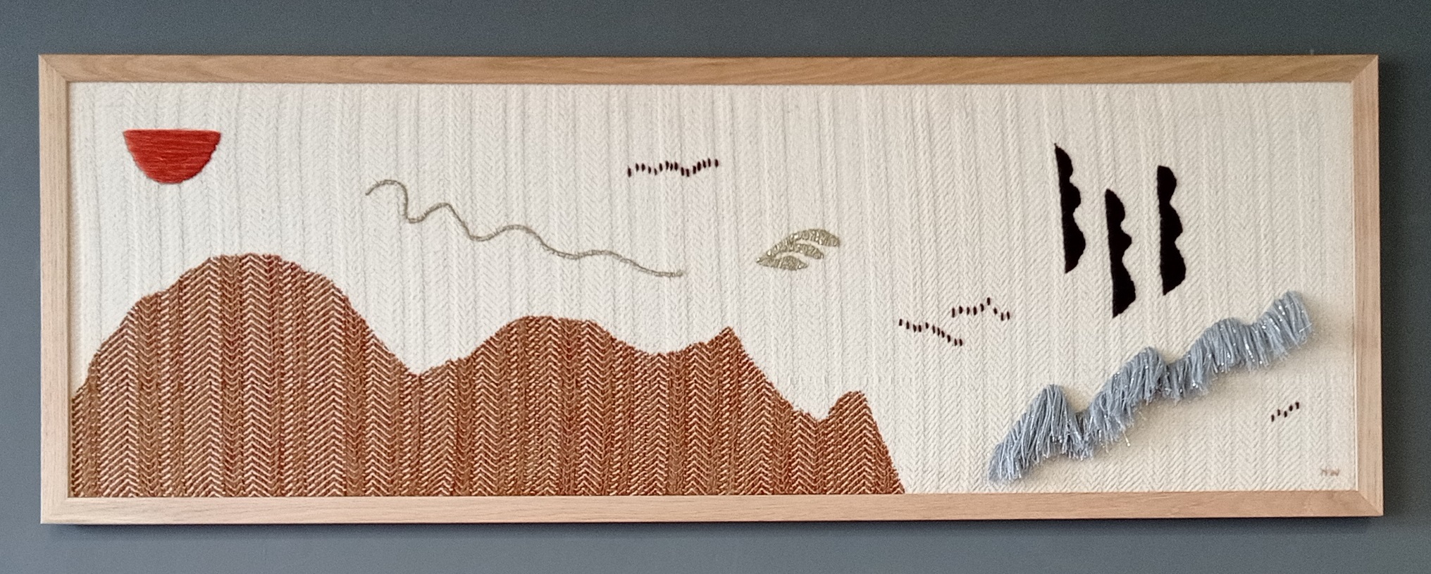 A figurative textile work. Brown shape reminiscent of a hill against an off-white background with features suggestive of natural formations. Framed in light wood and hanging on a grey wall.